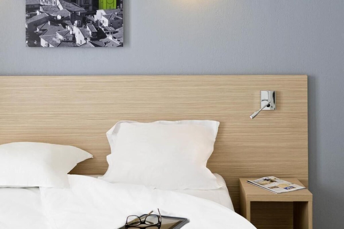 A neatly made bed with white bedding, a bedside table with a lamp, and a wall-mounted power outlet.