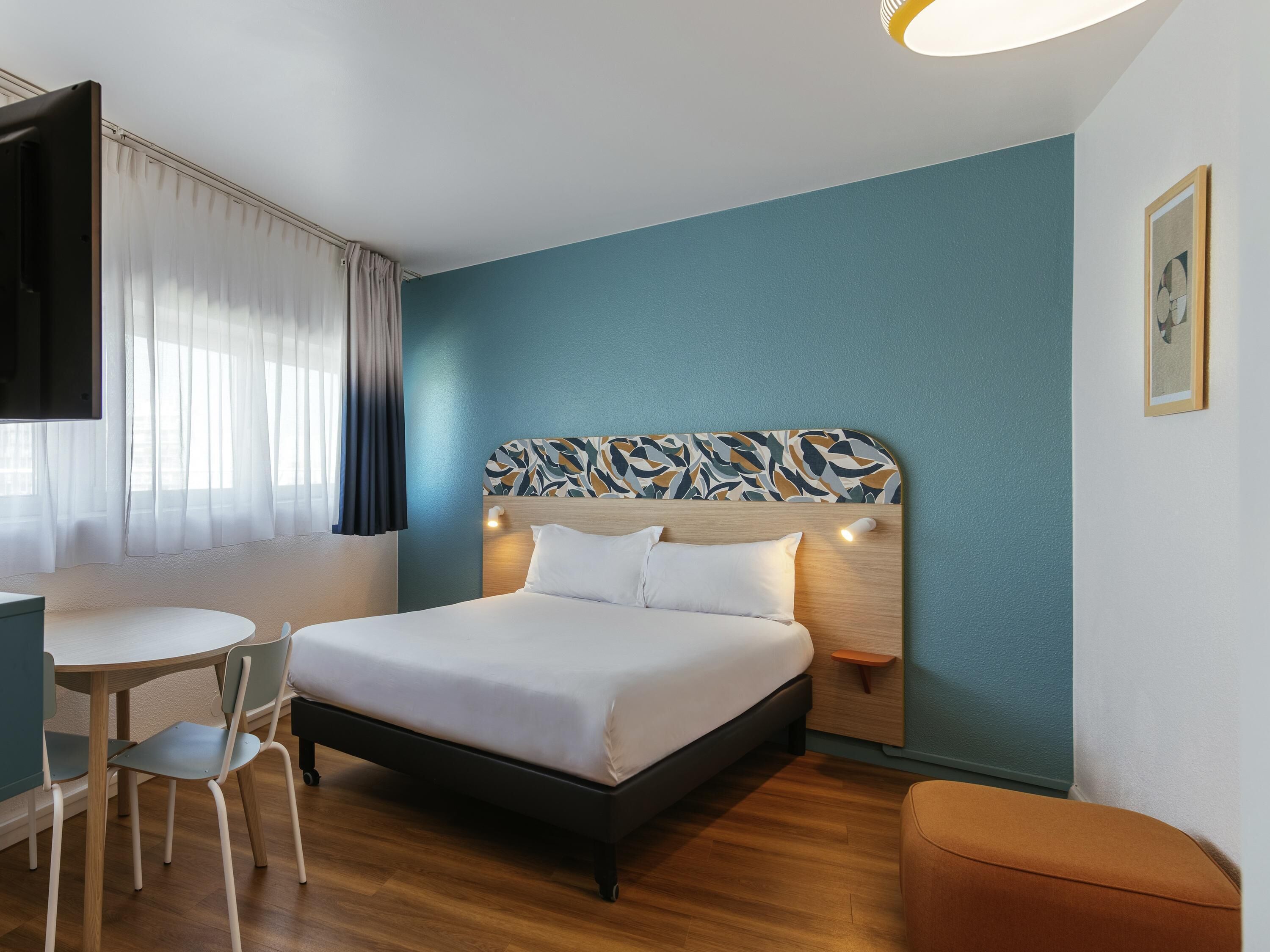 Quarto de hotel com cama, escrivaninha, cadeira, televisão e um quadro na parede.