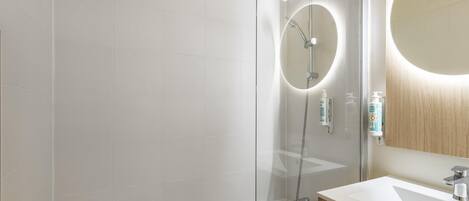 Un baño moderno con ducha envuelta en vidrio, tocador de madera y espejo redondo con luz.