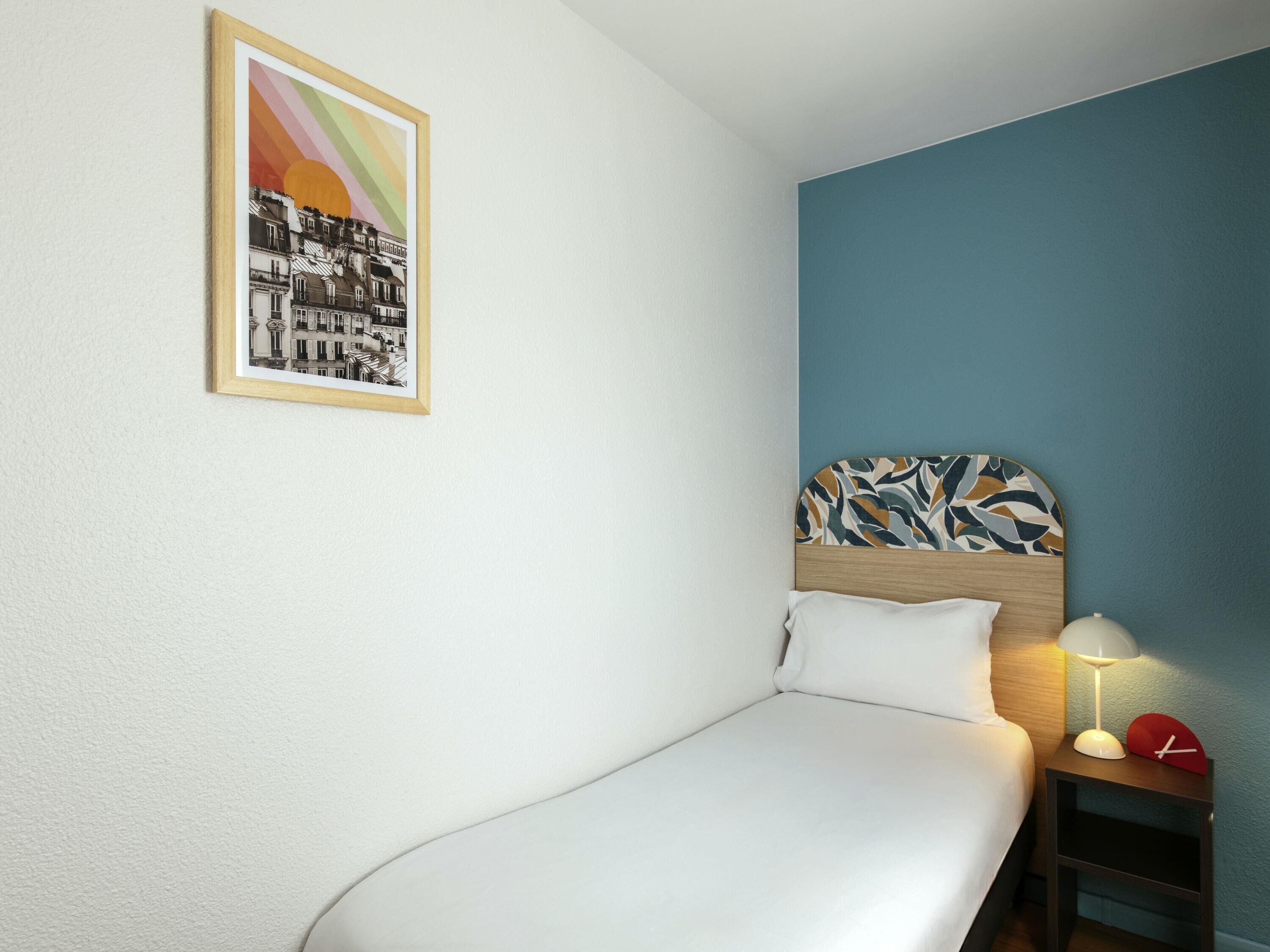 Un lit simple avec une tête de lit, une table de chevet avec une lampe, une horloge et un tableau encadré au mur.