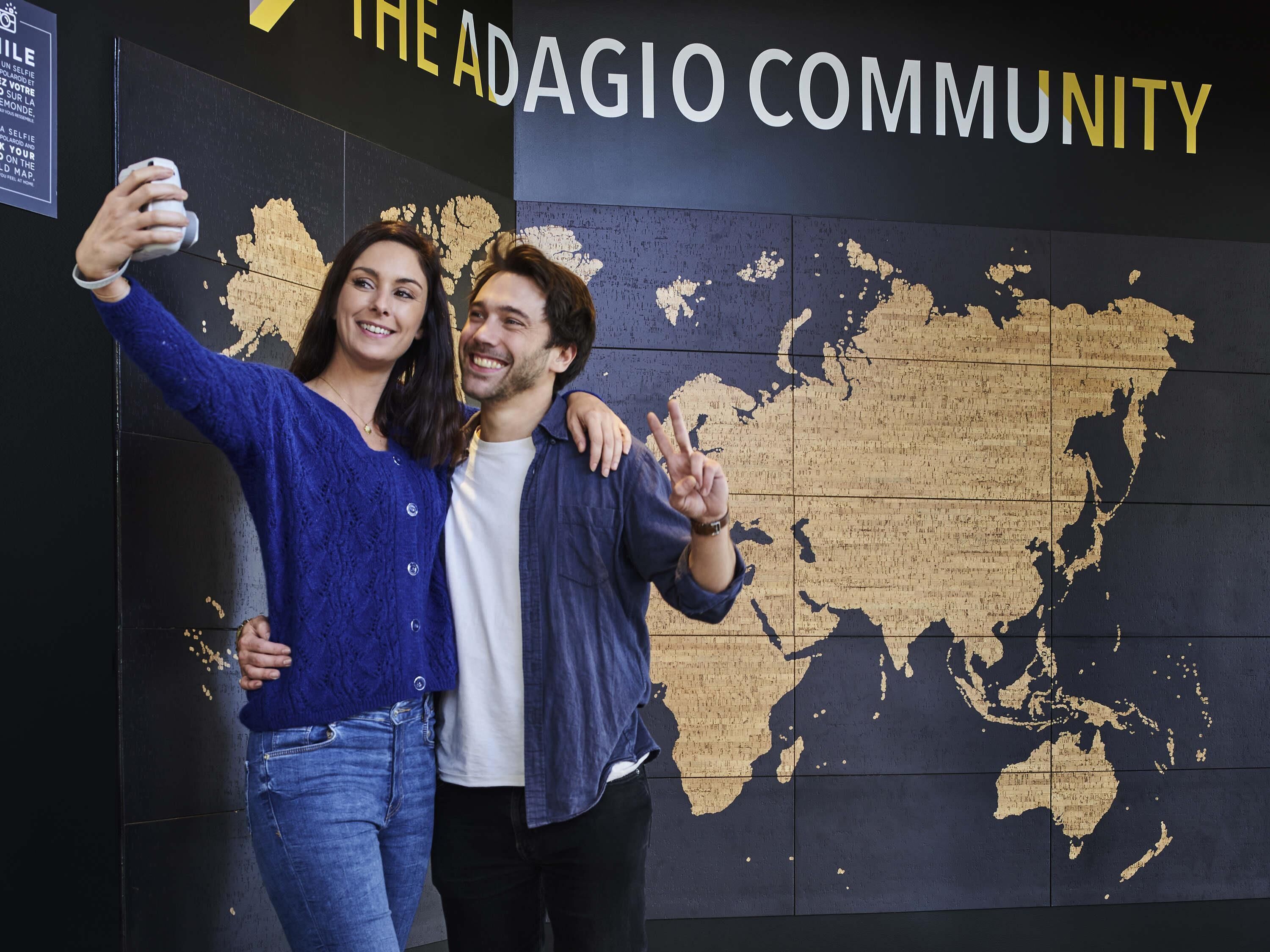 Two people taking a selfie in front of a wall with a map and the text 'THE ADAGIO COMMUNITY'.