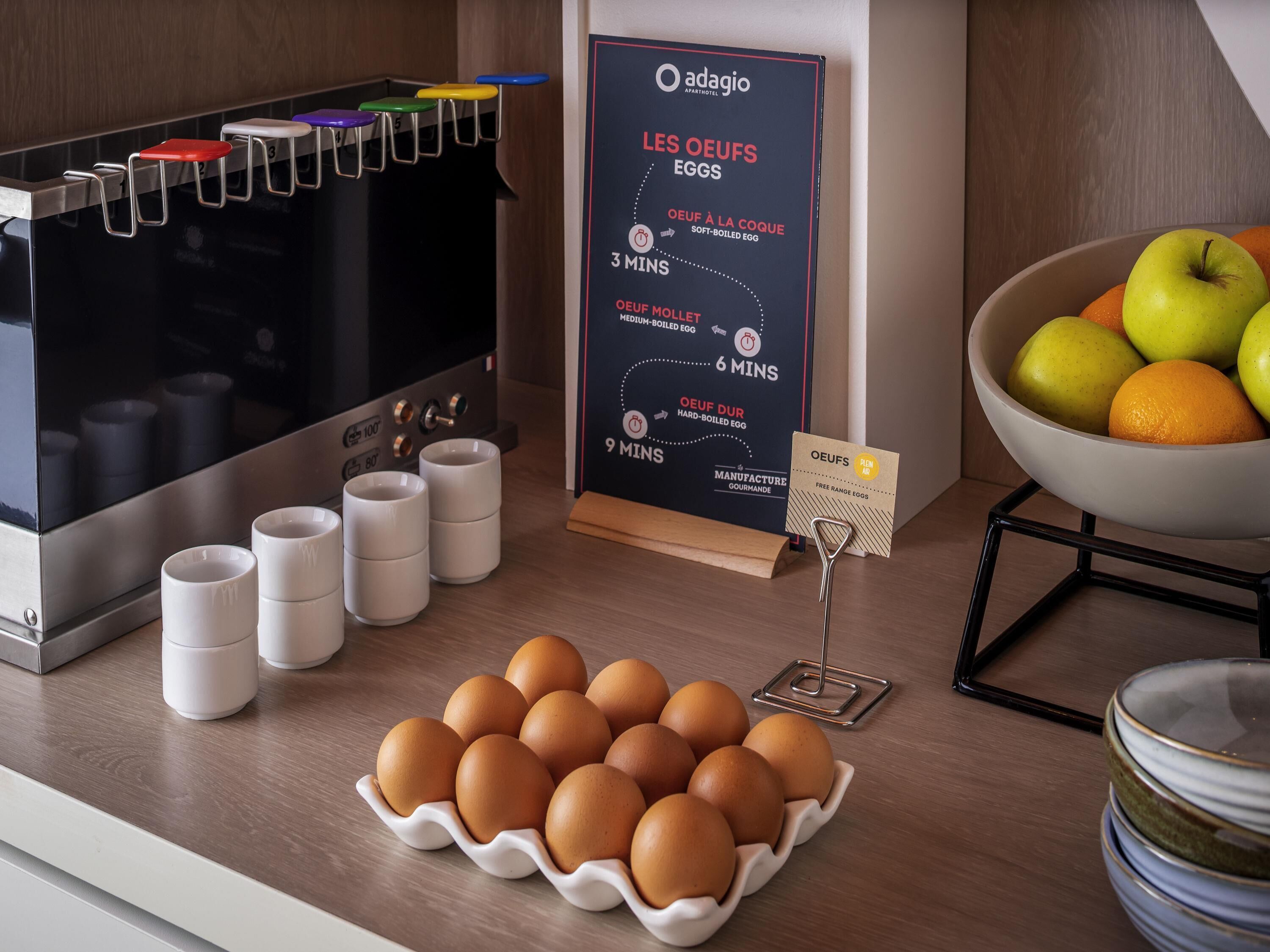 A breakfast station with a coffee machine, a poster detailing egg preparation times, and a bowl of fruit.