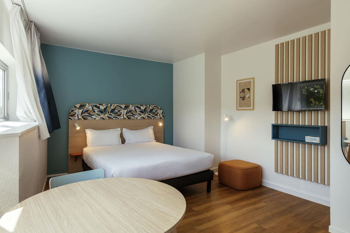 Ein Hotelzimmer mit Bett, rundem Tisch, an der Wand montiertem Fernseher und einem Fenster mit Vorhängen.