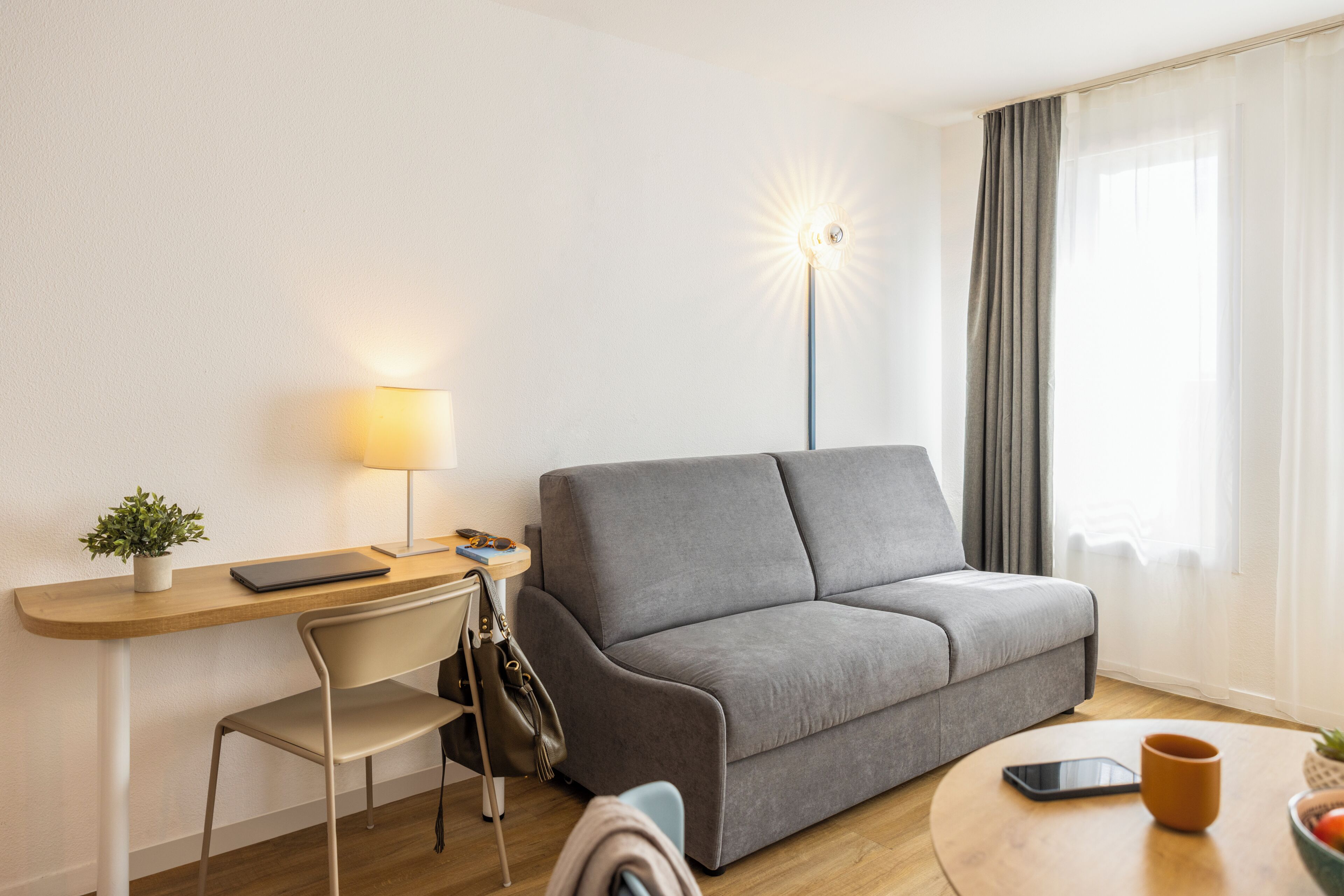 A modern living room with a sofa, a desk with a lamp, a chair, a smartphone, and a coffee cup.