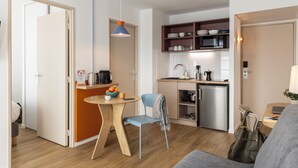 Una cocina compacta con microondas, pileta y alacenas, un comedor con mesa y sillas, y un sofá gris en un pequeño espacio de estar.