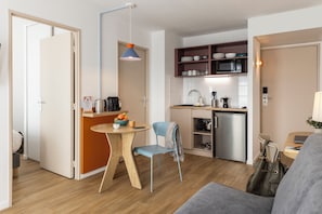 A compact kitchen with a microwave, sink, and cabinets, a dining area with a table and chairs, and a grey sofa in a small living space. - Aparthotel Adagio Access Toulouse Jolimont (Toulouse)