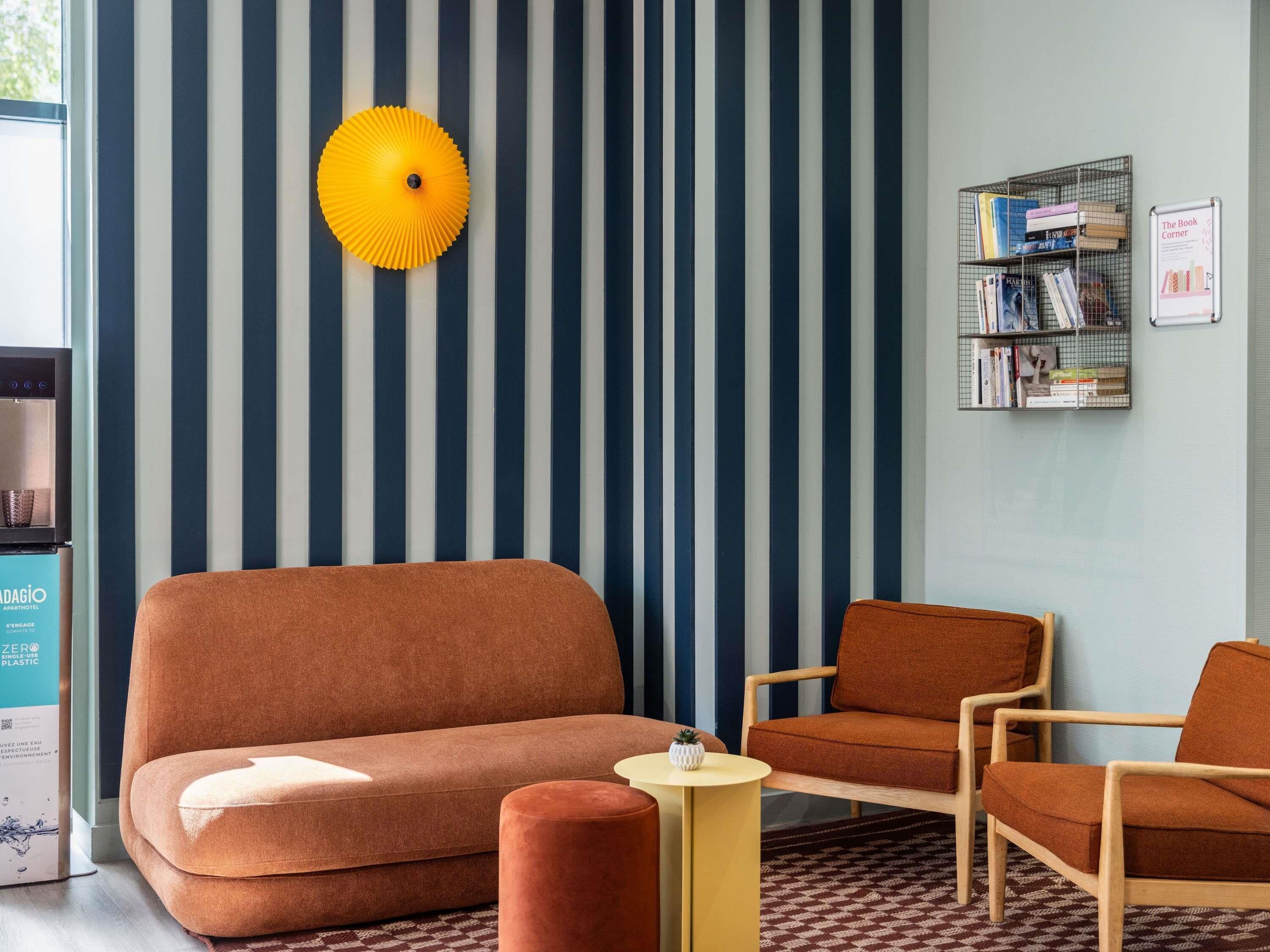 A living room with striped wallpaper, a sofa, two armchairs, a small round table, a yellow lamp, and a bookshelf.