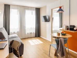 A modern hotel room with a sofa, a dining table with a bowl of fruit, a coffee machine, and a flat-screen TV. - Aparthotel Adagio Access Toulouse Jolimont (Toulouse)