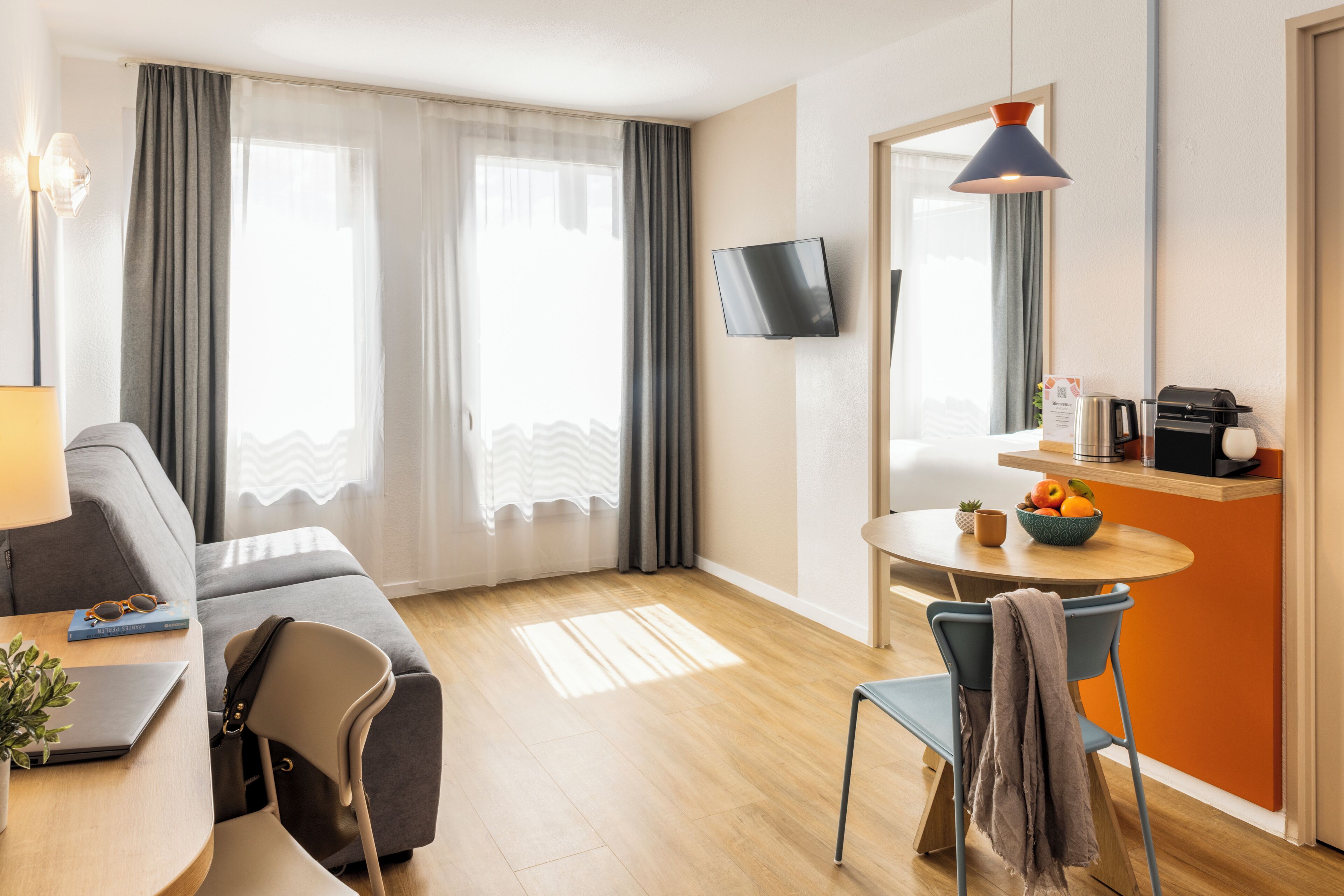 A modern hotel room with a sofa, dining table, chairs, a TV, and a small kitchen area with a coffee machine.