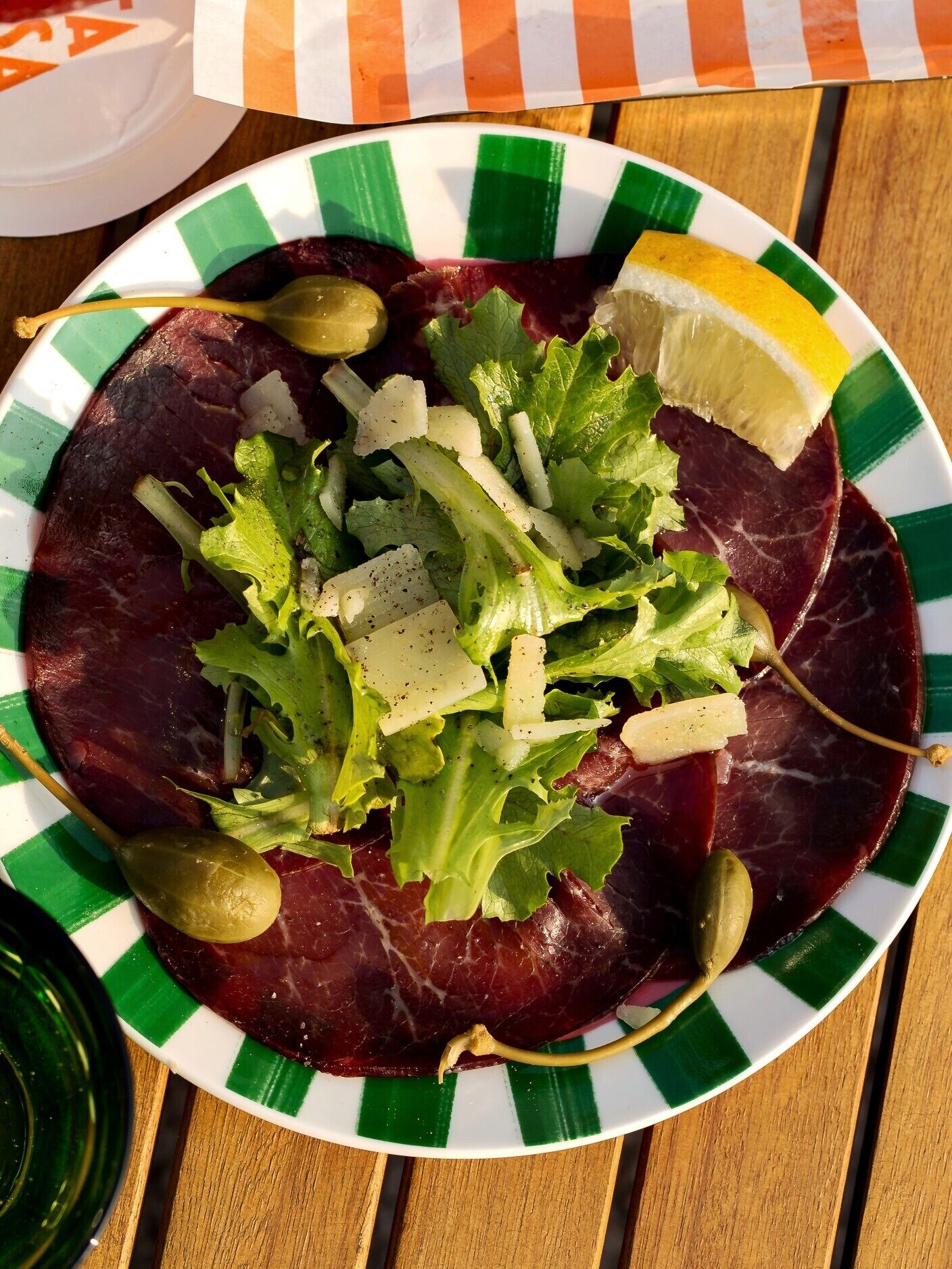 Un plato de comida con un borde verde y blanco a rayas, que contiene carne oscura, verduras de hoja verde y un gajo de limón.