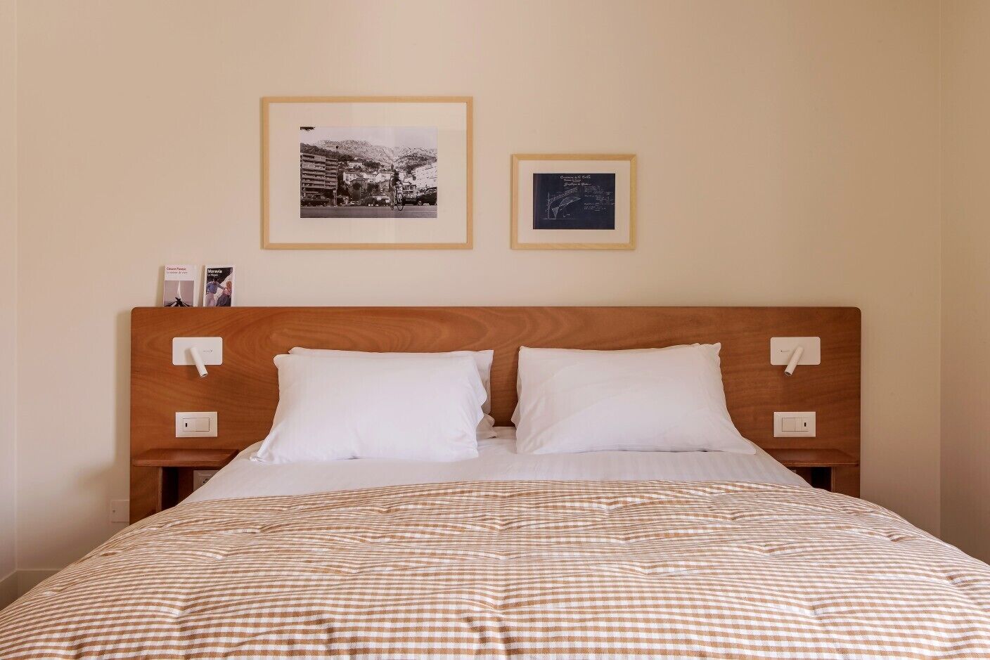 Una cama bien hecha con una colcha a cuadros, dos almohadas blancas y un cabecero de madera. Se ven dos interruptores de luz en la pared y dos cuadros enmarcados sobre la cama.