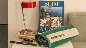 Quarto de hotel bem arrumado com um quadro na parede, uma luminária vermelha, um livro intitulado 'SLIM AARON' e uma toalha verde com 'ALFRED HOTELS'.
