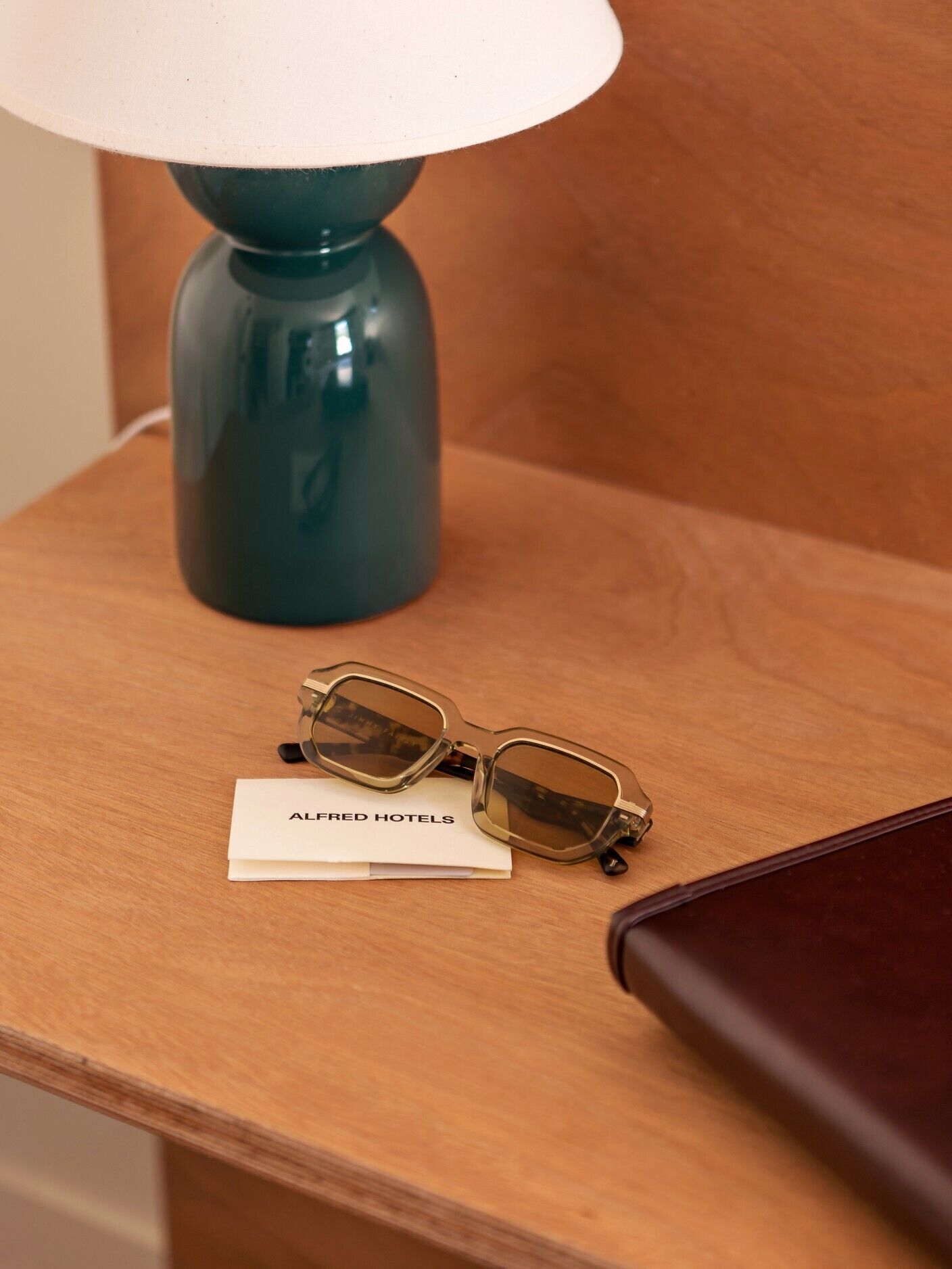 A wooden nightstand with a teal lamp, a pair of glasses, and a white card with "ALFRED HOTELS" text.
