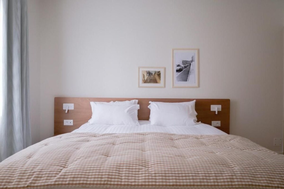 Un lit bien fait, recouvert d’une courtepointe à carreaux, avec deux oreillers blancs et une tête de lit en bois. Deux tableaux encadrés sont accrochés au mur, au-dessus du lit.