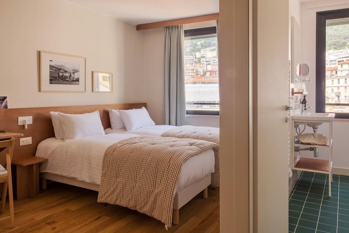 Habitación de hotel con dos camas, un escritorio, una silla, una ventana con vistas a edificios y un baño con lavamanos y espejo.
