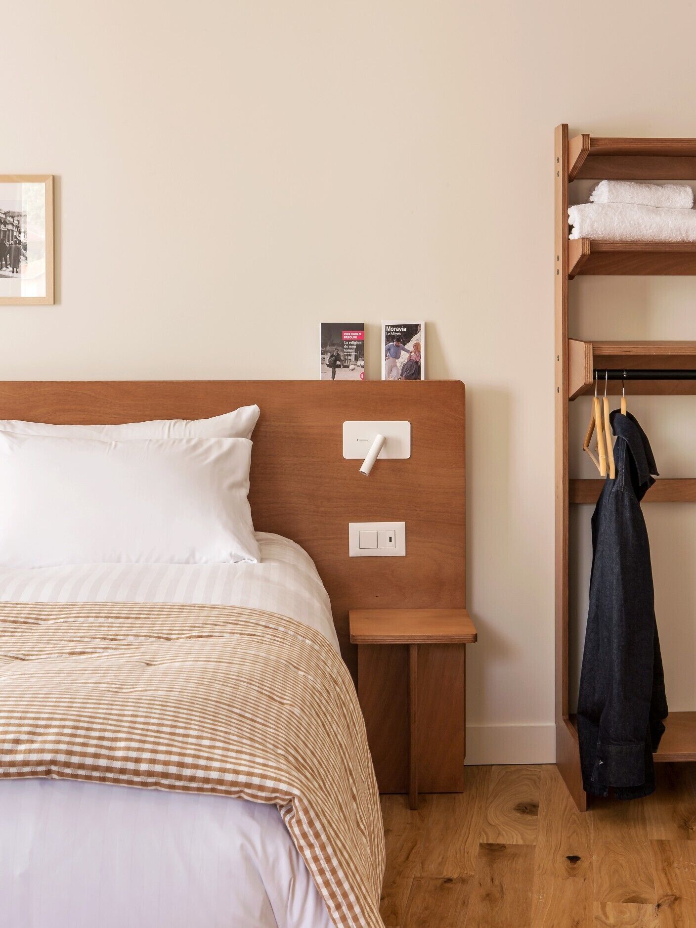 A neatly made bed with a checkered blanket, a bedside table with a light switch, a small shelf with books, and a wooden wardrobe with hanging clothes.