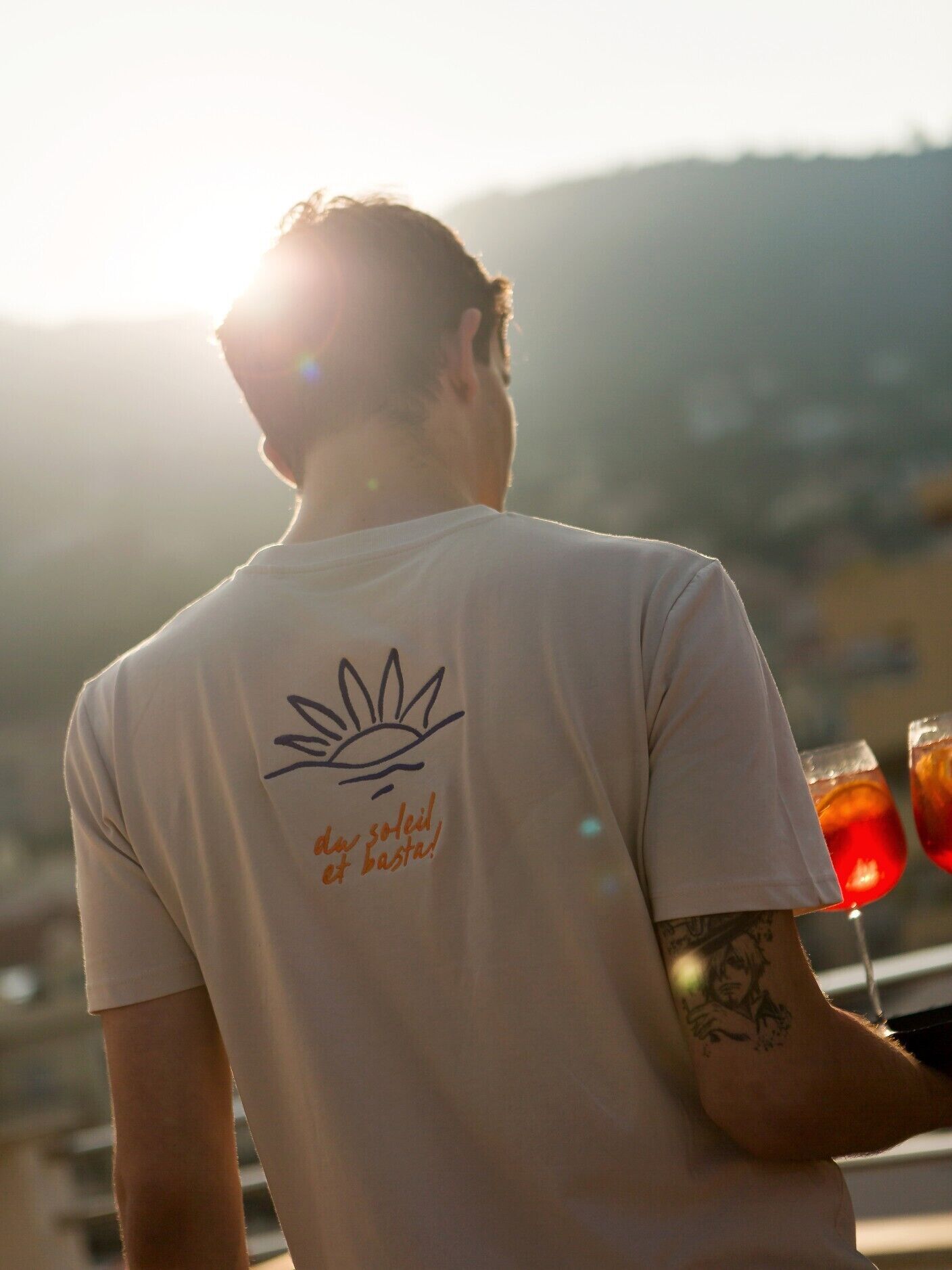 A person wearing a white t-shirt with a sun and text on the back, standing with a tattooed arm, and holding a drink.