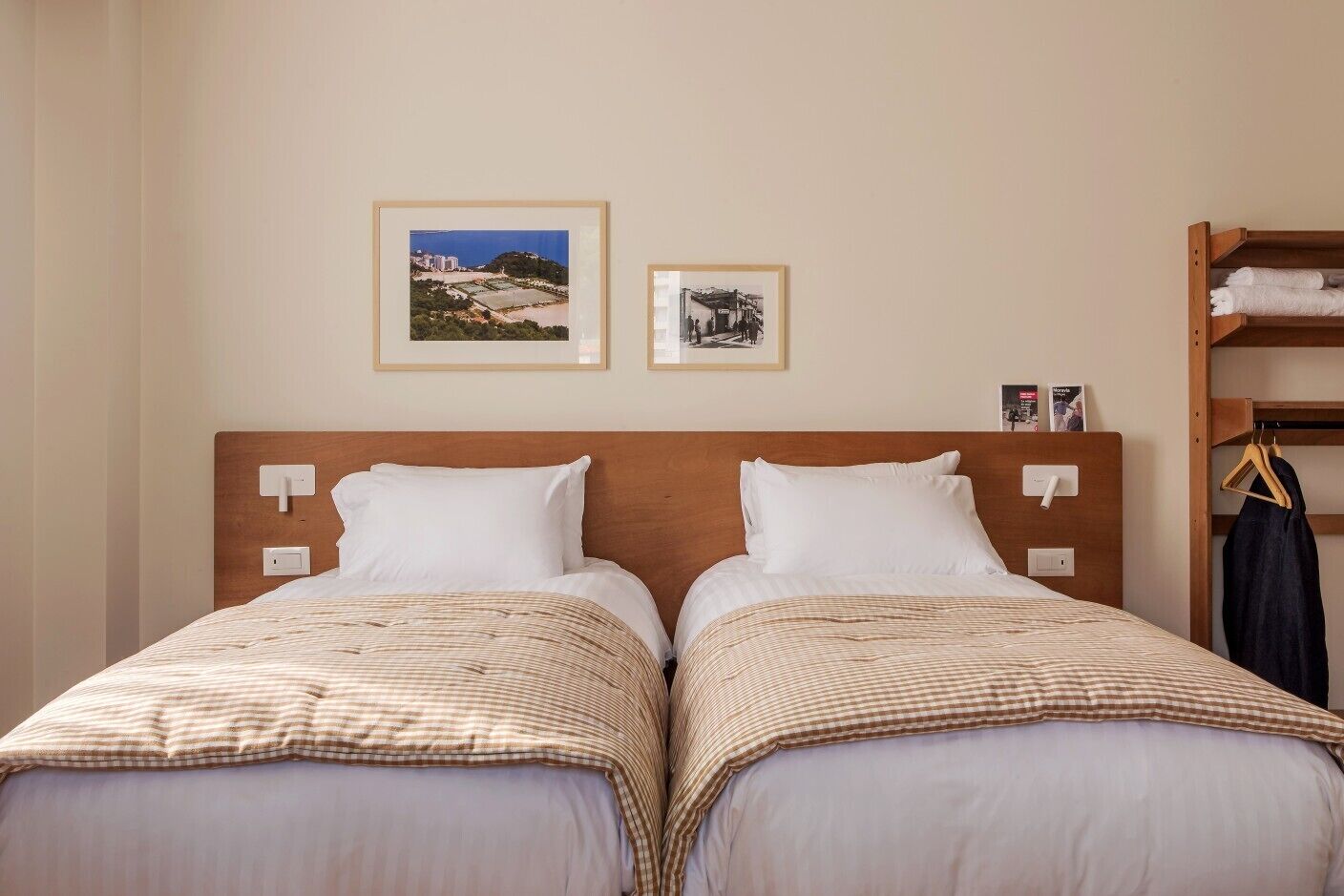 Ein Hotelzimmer mit zwei Betten, einem Holzkopfteil, einem Regal mit Handtüchern und zwei gerahmten Bildern an der Wand.