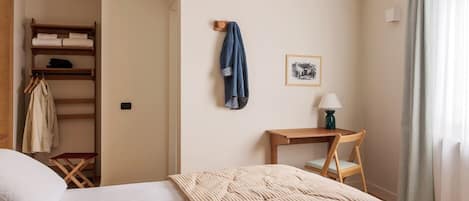 A hotel room with a bed, a wooden chair, a desk with a lamp, a coat rack, and a wardrobe.