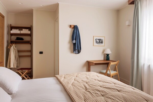 A hotel room with a bed, a wooden chair, a desk with a lamp, a coat rack, and a wardrobe.