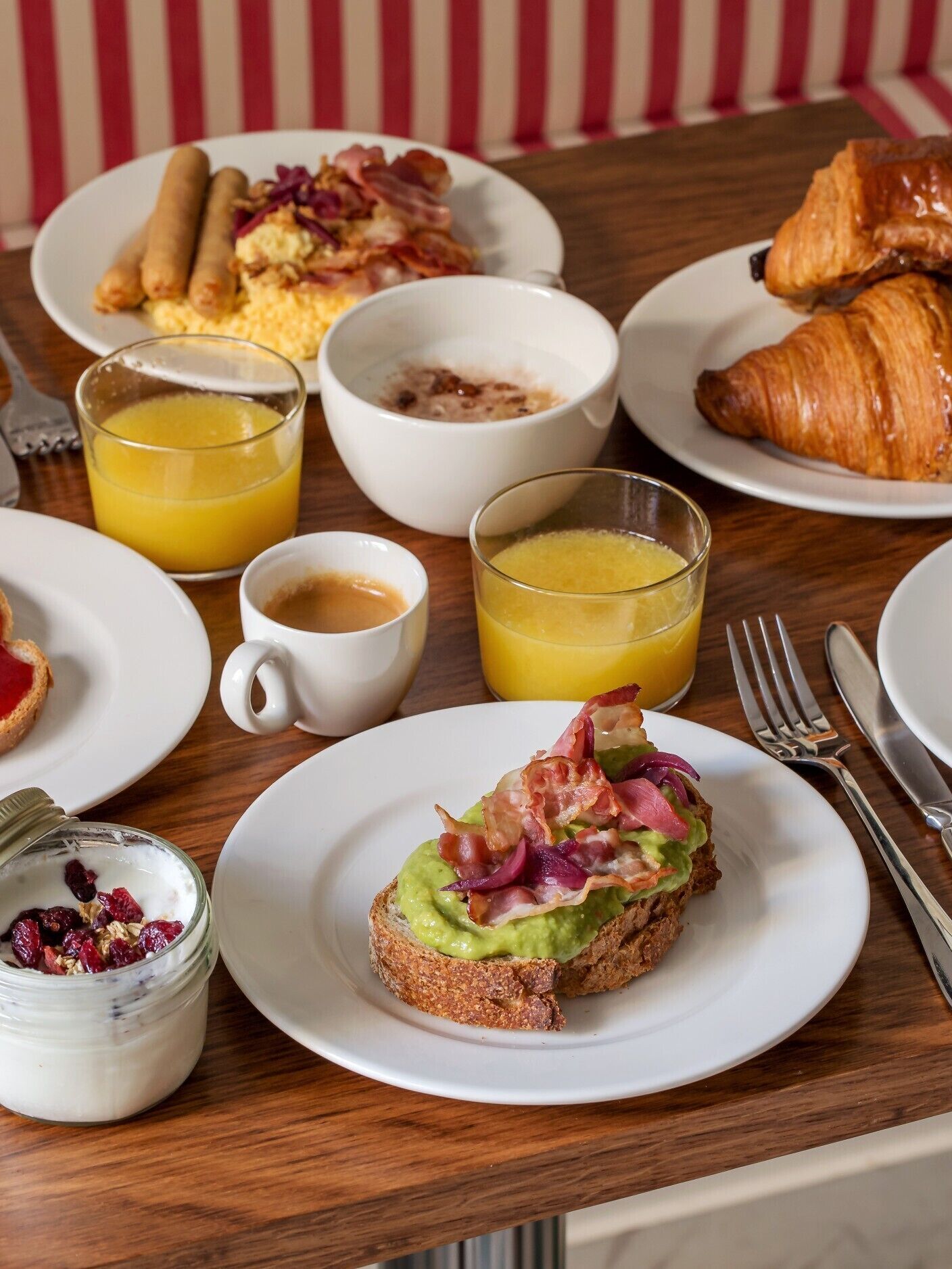 Eixam de matí amb plats de menjar, incloent torrades amb avocado i bacon, ous barrejats, botifarreta i un croissant. També hi ha gots de suc d'aranja i un bol petit de iogurt amb baies.