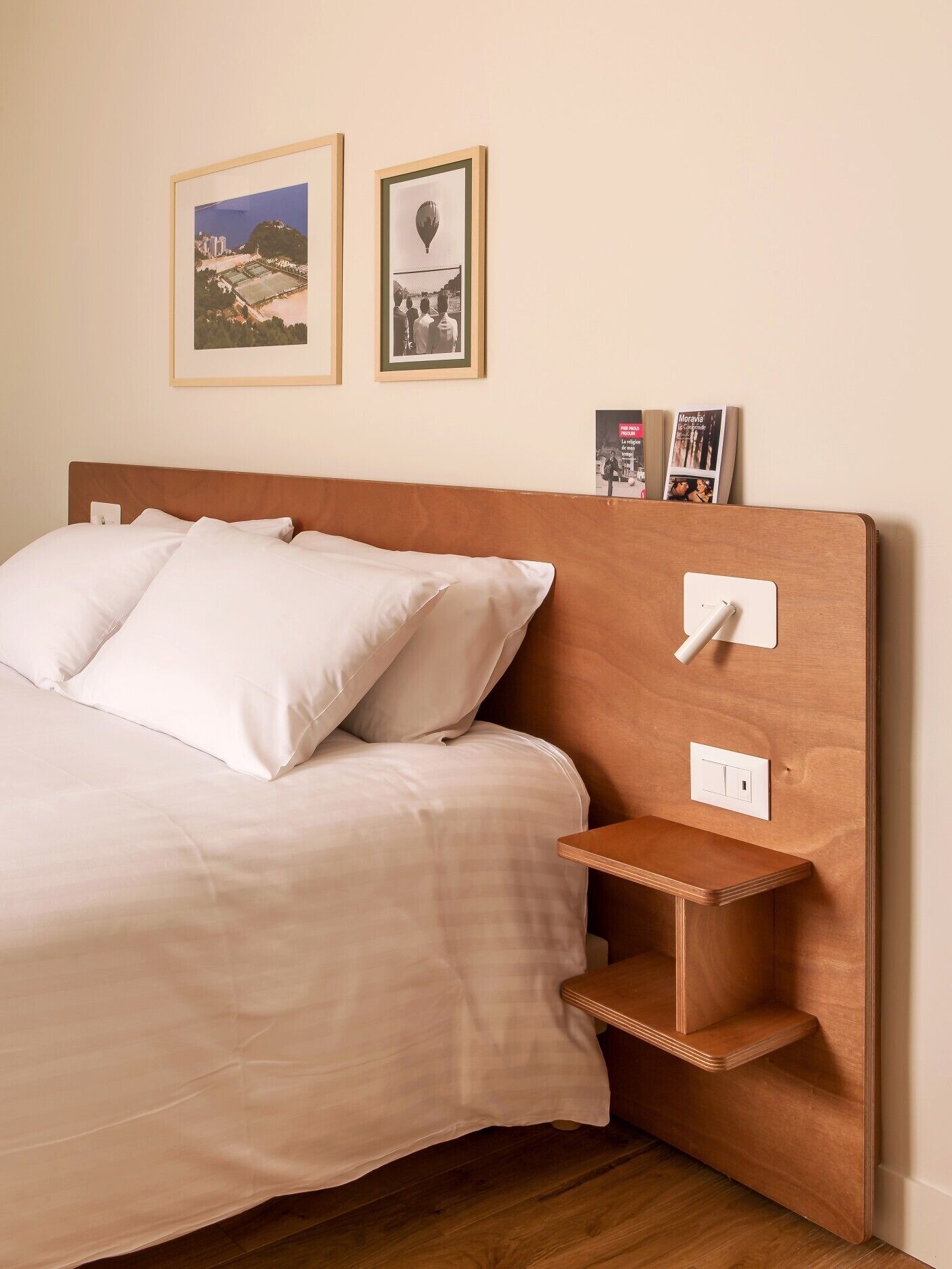 A neatly made bed with white linens, a wooden headboard, and a small wooden nightstand with a light switch.