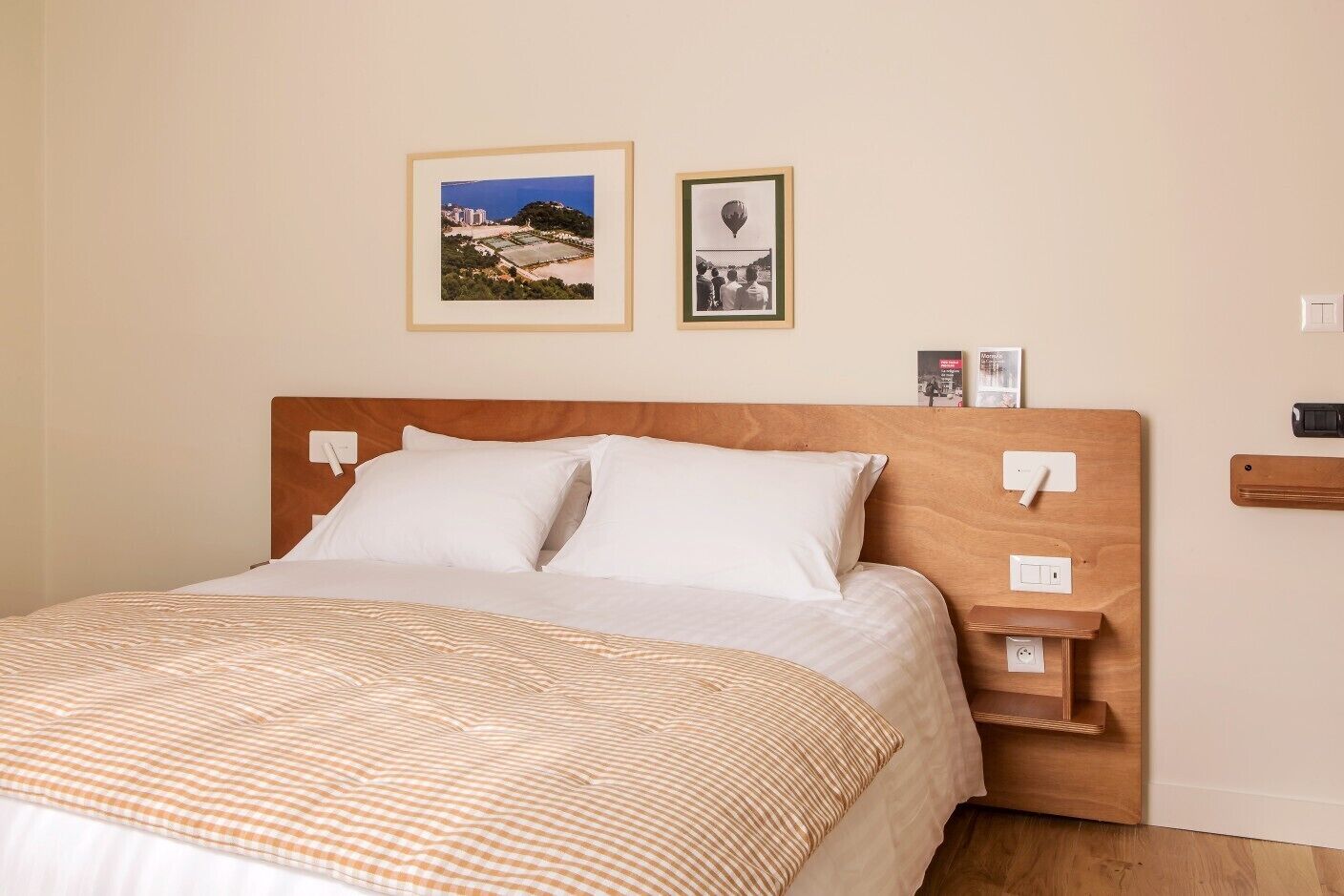 Een hotelkamer met een houten hoofdeinde, een bed met witte lakens, een geruit plaid en twee ingelijste foto's aan de muur.
