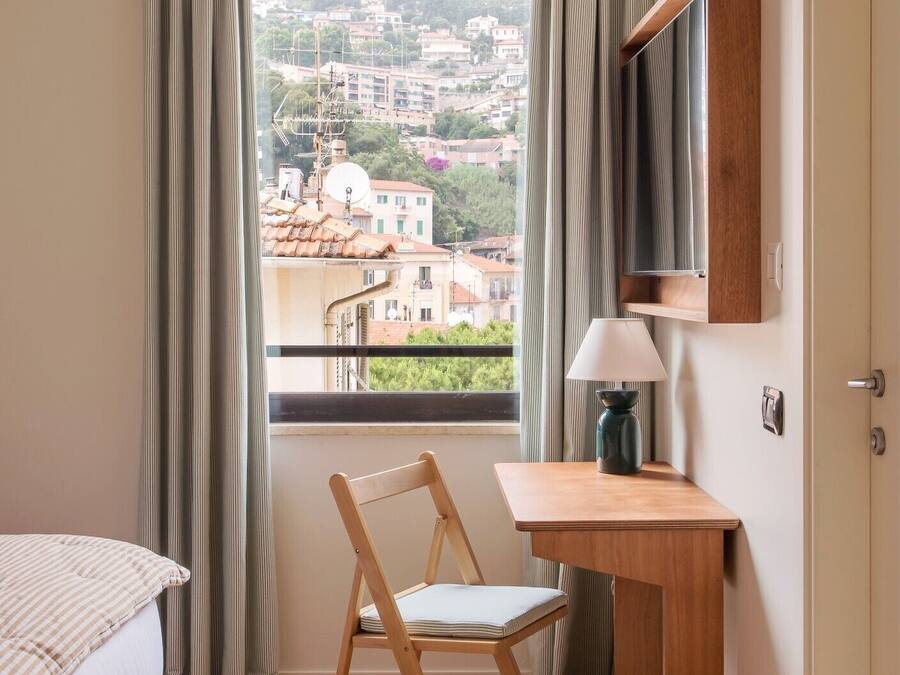 A small, well-lit room with a wooden desk, a chair, and a bed. A window offers a view of a cityscape.