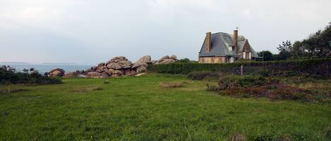 Uma casa com fachada de pedra e telhado inclinado, cercada por um campo gramado e pedras grandes.
