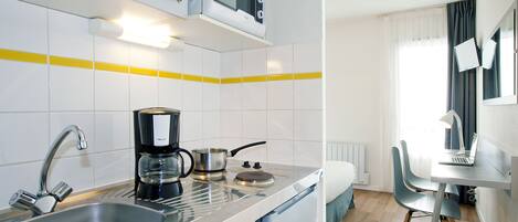A compact kitchen with a microwave, coffee maker, and kettle on the counter, a sink, and a small dining area with a chair and table.