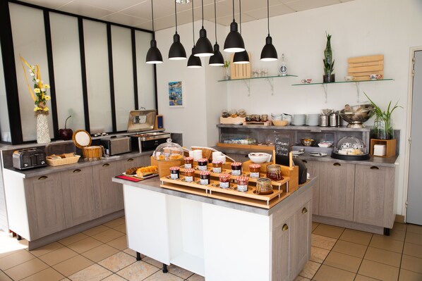 A modern kitchen with a central island displaying various food items, a coffee machine, and a microwave. There are hanging lights, shelves with decorative items, and a large window providing natural light.