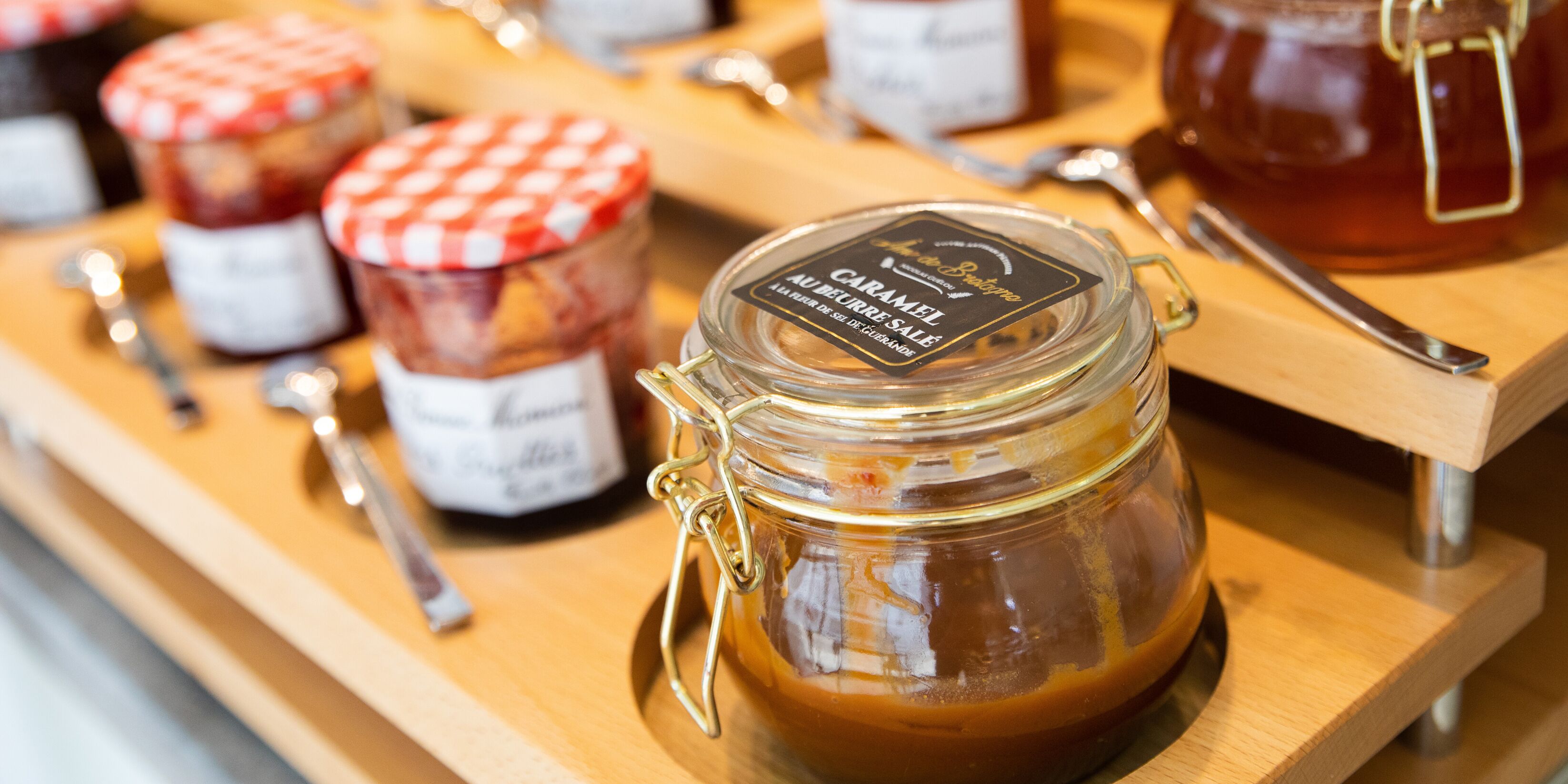 Exhibición de tarros de salsa de caramelo con tapas y asas de metal, dispuestos sobre una tabla de madera con cucharas a un lado.