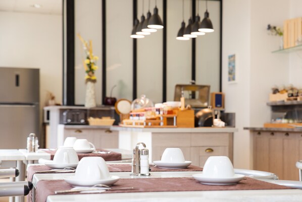 A modern dining area with tables set for service, a kitchen in the background, and pendant lighting. - BRIT HOTEL Confort Lannion Centre Gare (Lannion)