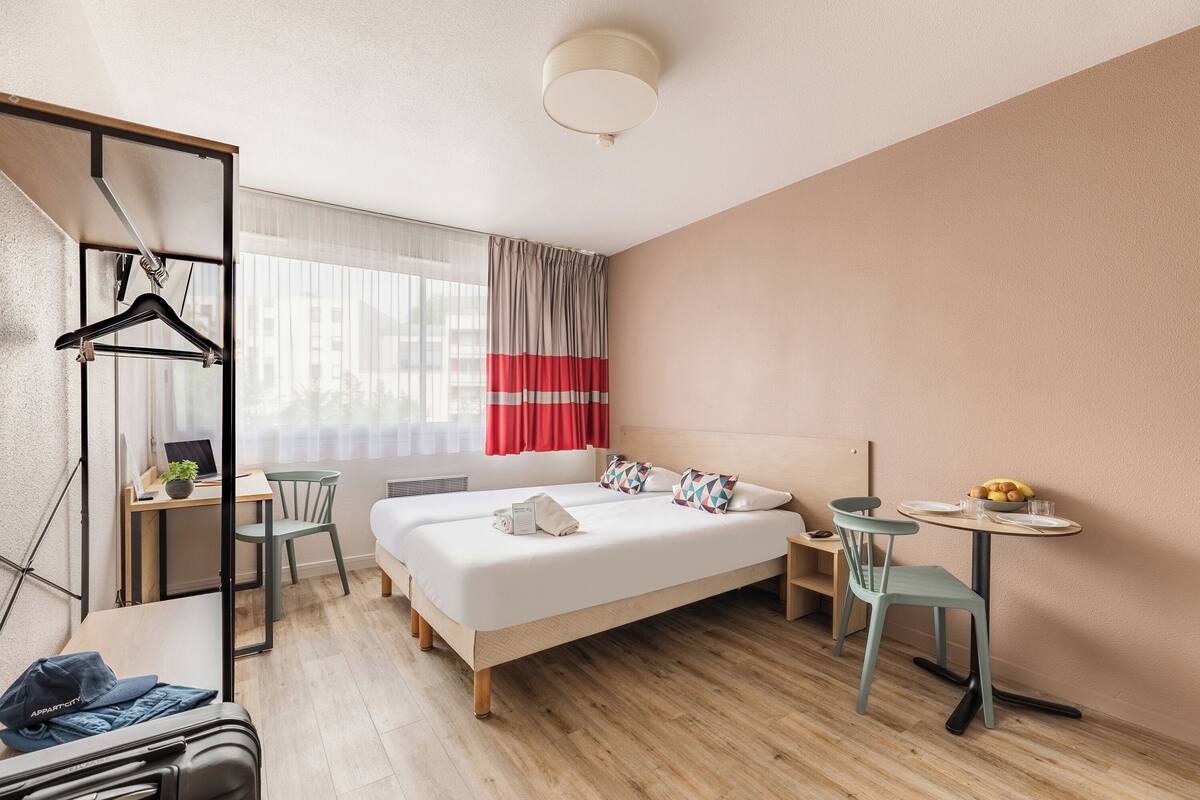 A modern hotel room with a bed, a desk, a chair, and a small table with a fruit bowl.