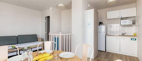 A modern, well-lit room with a dining table set for two, a sofa, and a kitchenette with white cabinets and a microwave.