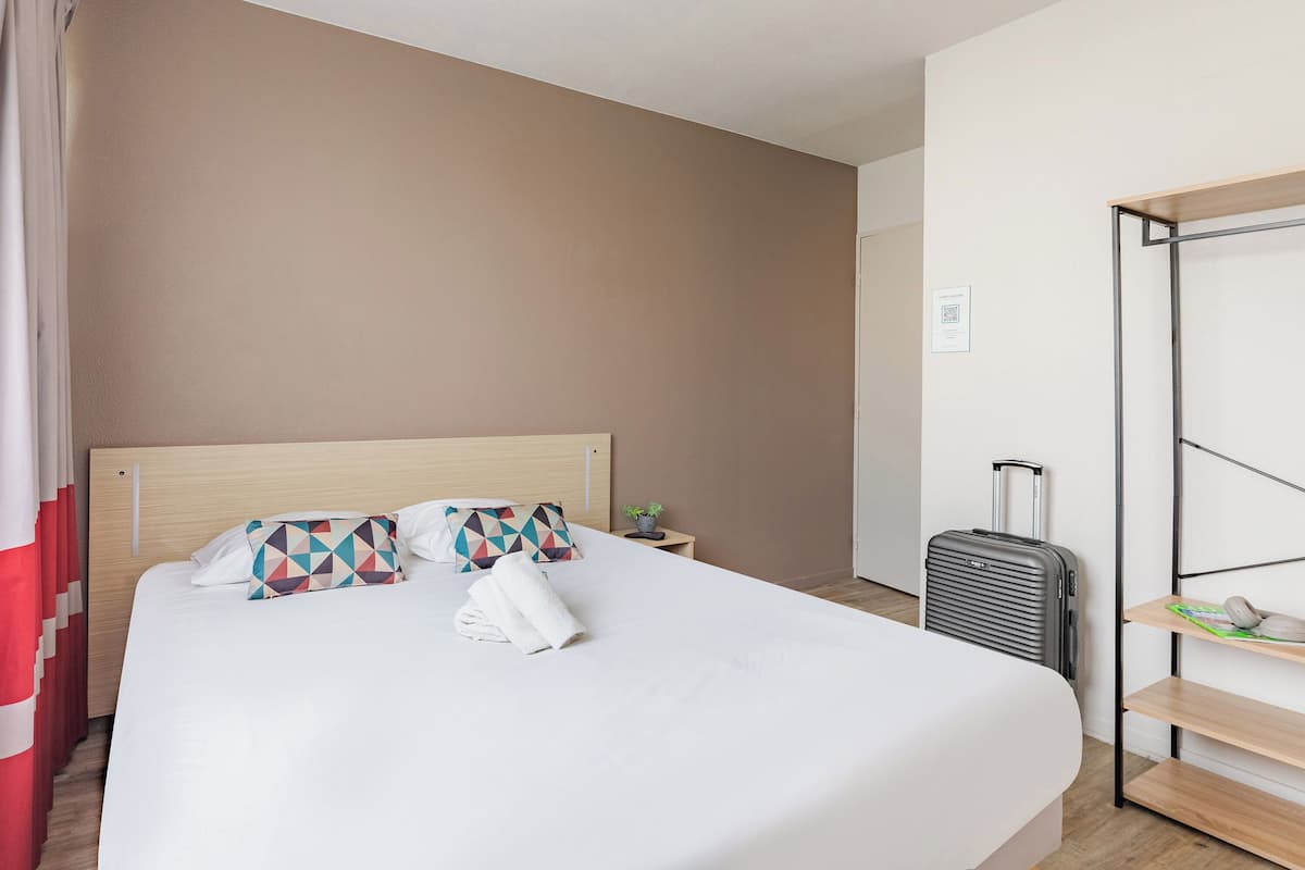 A neatly made bed with pillows, a small bedside table with a plant, and a suitcase against a wall.