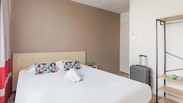 Una cama bien hecha con almohadas, una mesita de noche con una planta y una maleta apoyada en la pared.