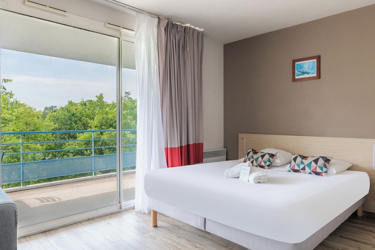 A hotel room with a large bed, a view of greenery through the balcony, and a framed picture on the wall.
