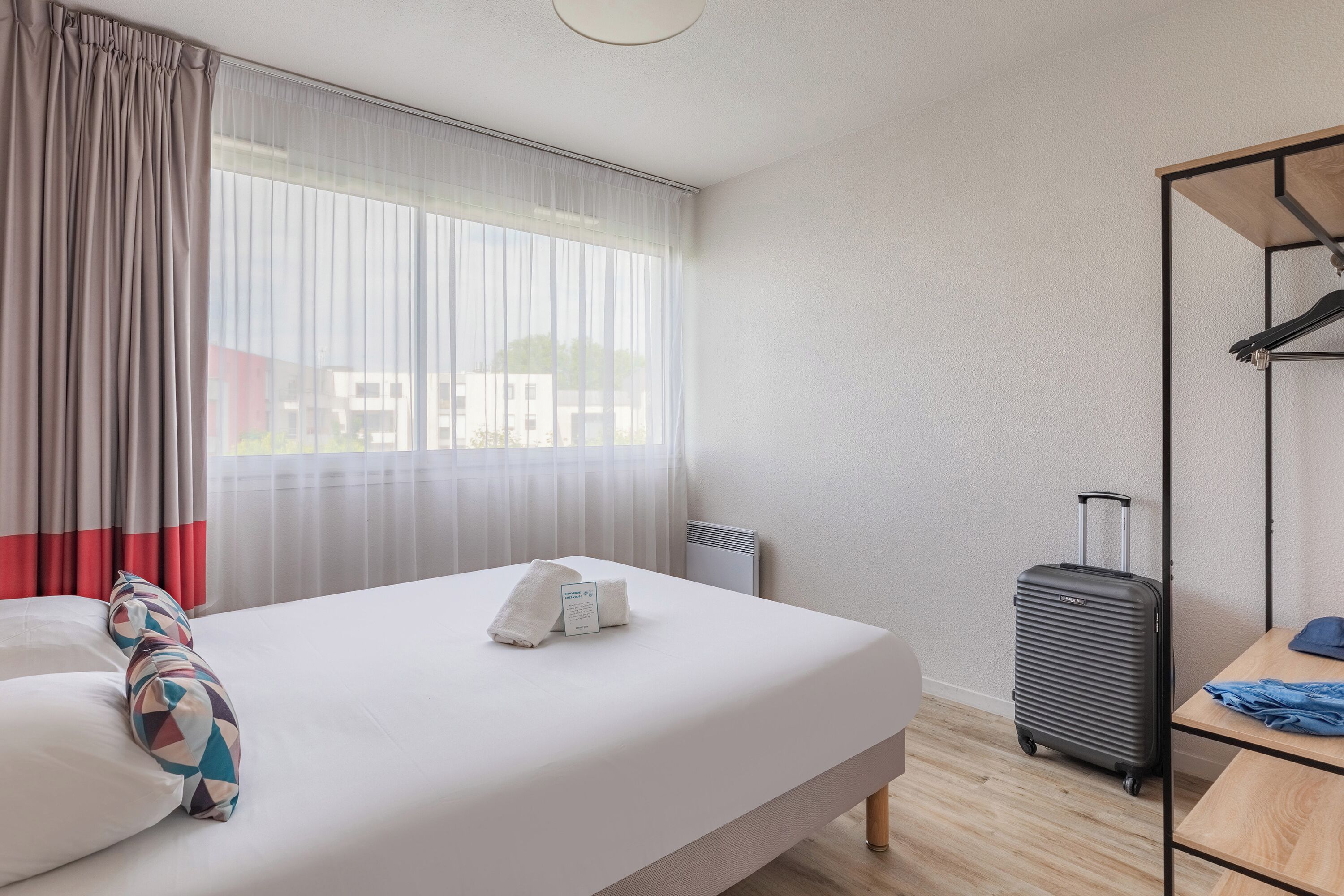 Une chambre d’hôtel avec un lit, une valise, une table et une fenêtre avec des rideaux.