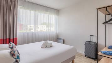 Une chambre d’hôtel avec un lit, une valise, une table et une fenêtre avec des rideaux.