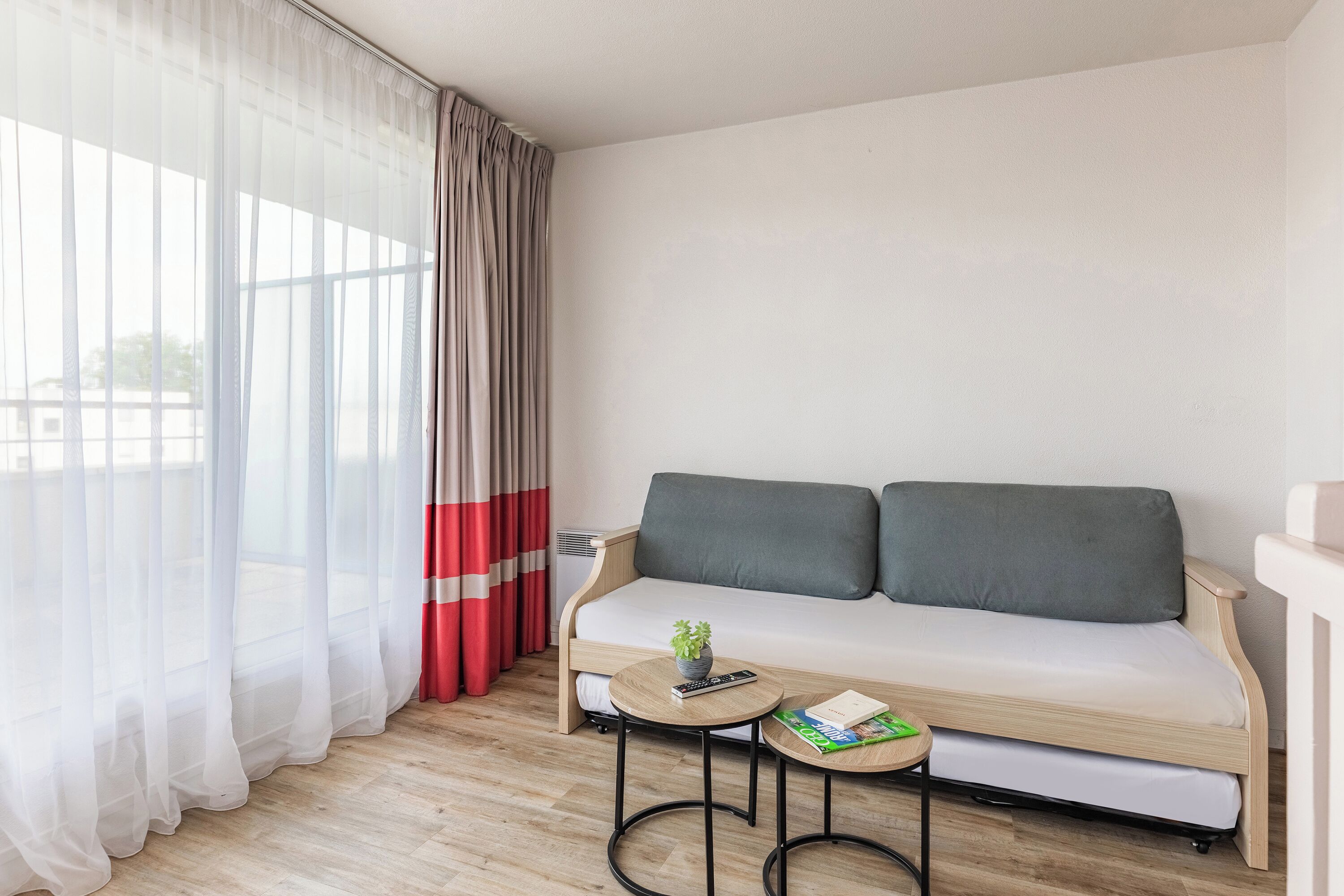 A modern room with a sofa, two small tables, a plant, and a window with sheer curtains.