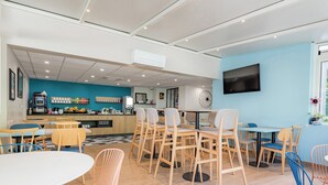 A modern cafe with wooden tables and chairs, a blue accent wall, and a flat-screen TV. - Appart'City Classic La Rochelle Centre (La Rochelle)