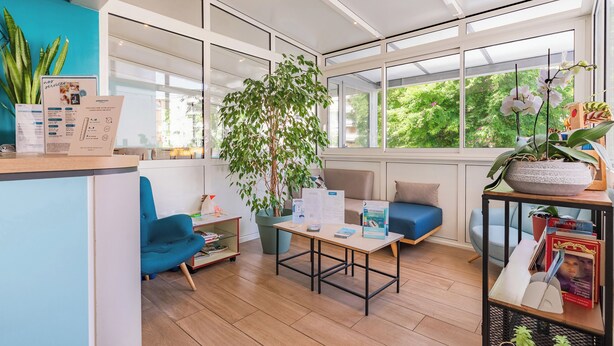 A modern reception area with a blue chair, a wooden table, a large potted plant, and a view of greenery outside. - Appart'City Classic La Rochelle Centre (La Rochelle)