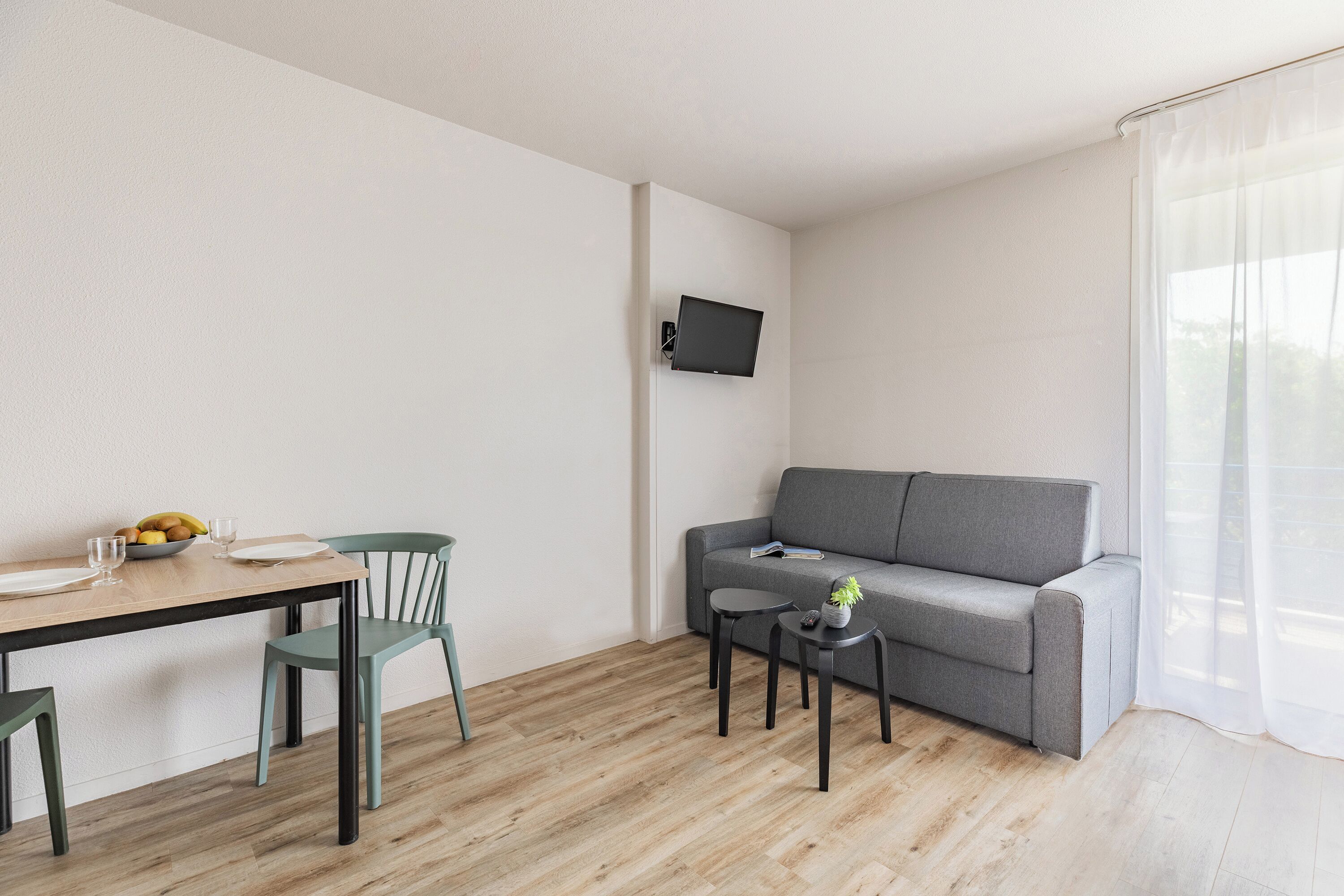 A modern living room with a grey sofa, a wooden dining table with chairs, a small side table, and a mounted TV.