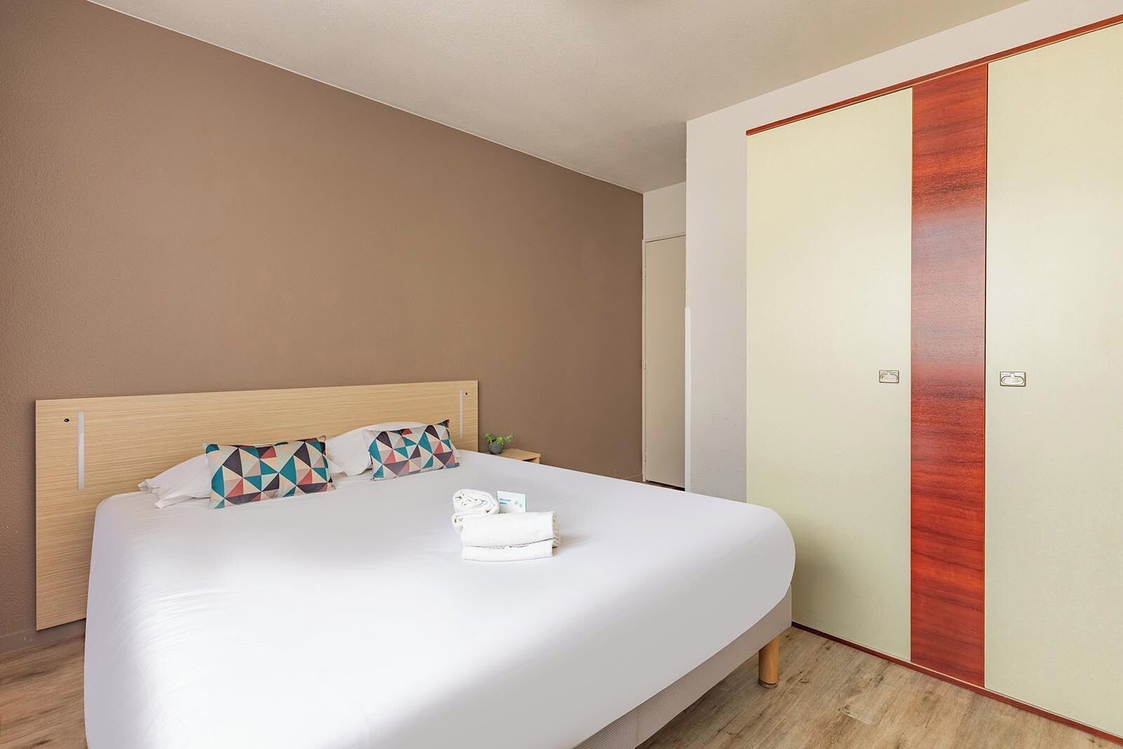Una cama bien tendida con un cabecero de madera, un almohadón con un diseño geométrico y una toalla doblada sobre la cama.