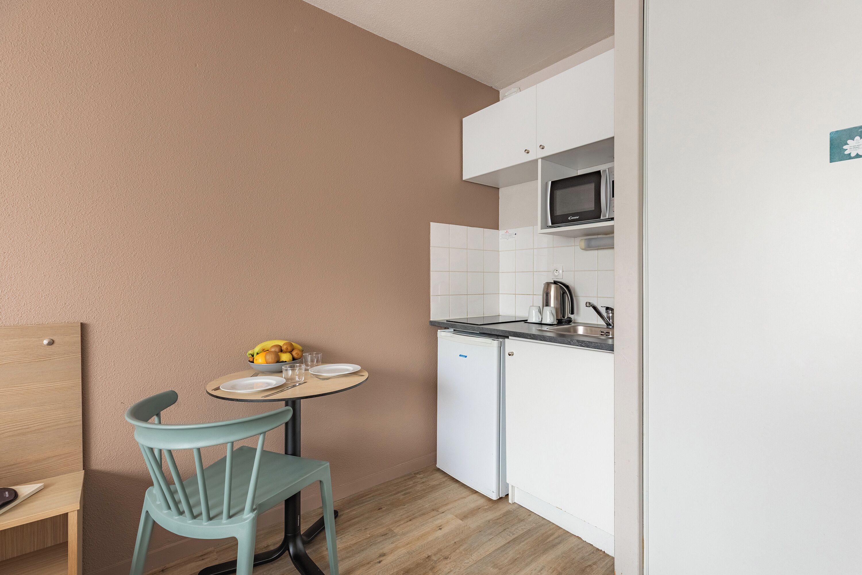 Cocina compacta con armarios blancos, un microondas y un lavamanos. Hay una mesa redonda pequeña con una silla, un cuenco de fruta y un plato.