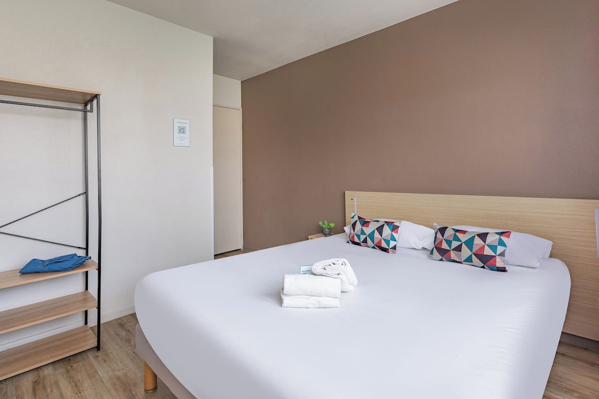 A hotel room with a large bed, a wooden shelf unit, and a small plant.