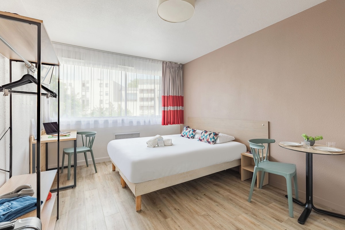 A modern hotel room with a large bed, a desk, a chair, a small table, and a window with curtains.