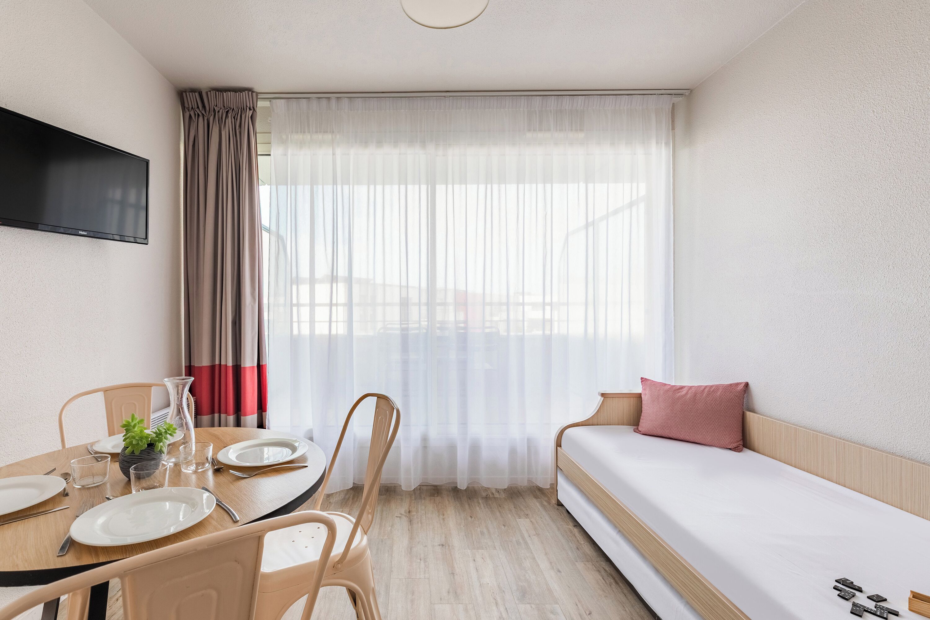 Een kleine hotelkamer met een bed, een eettafel met stoelen, een aan de muur bevestigde televisie en een raam met dunne gordijnen.