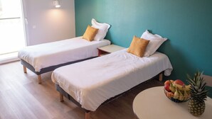 Dos camas individuales con sábanas blancas y almohadas marrones, una mesita con un frutero, y una pared color aguamarina.